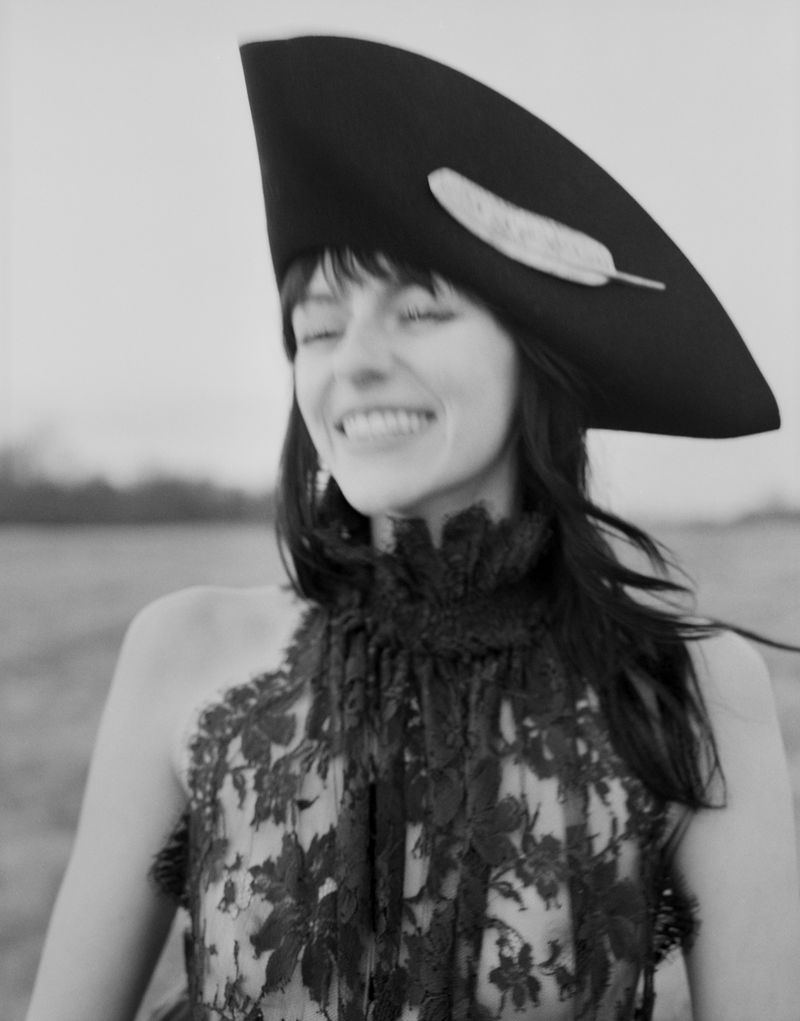 Monochrome portrait of smiling adult woman with closed eyes, wearing a large decorative hat with feather, and a lace top outdoors.
