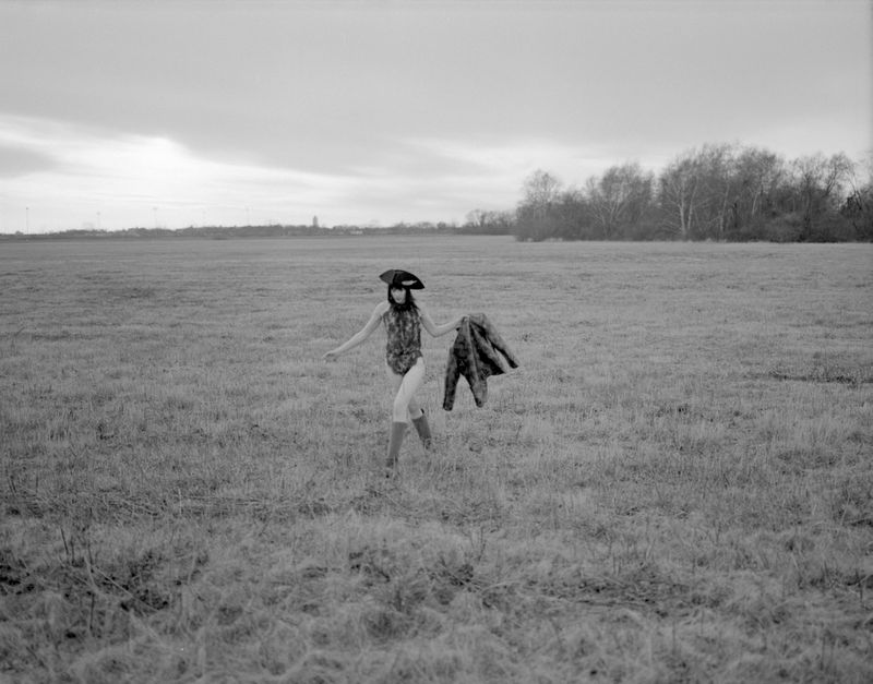Monochrome landscape editorial showing a figure walking across a grassy field carrying a patterned jacket