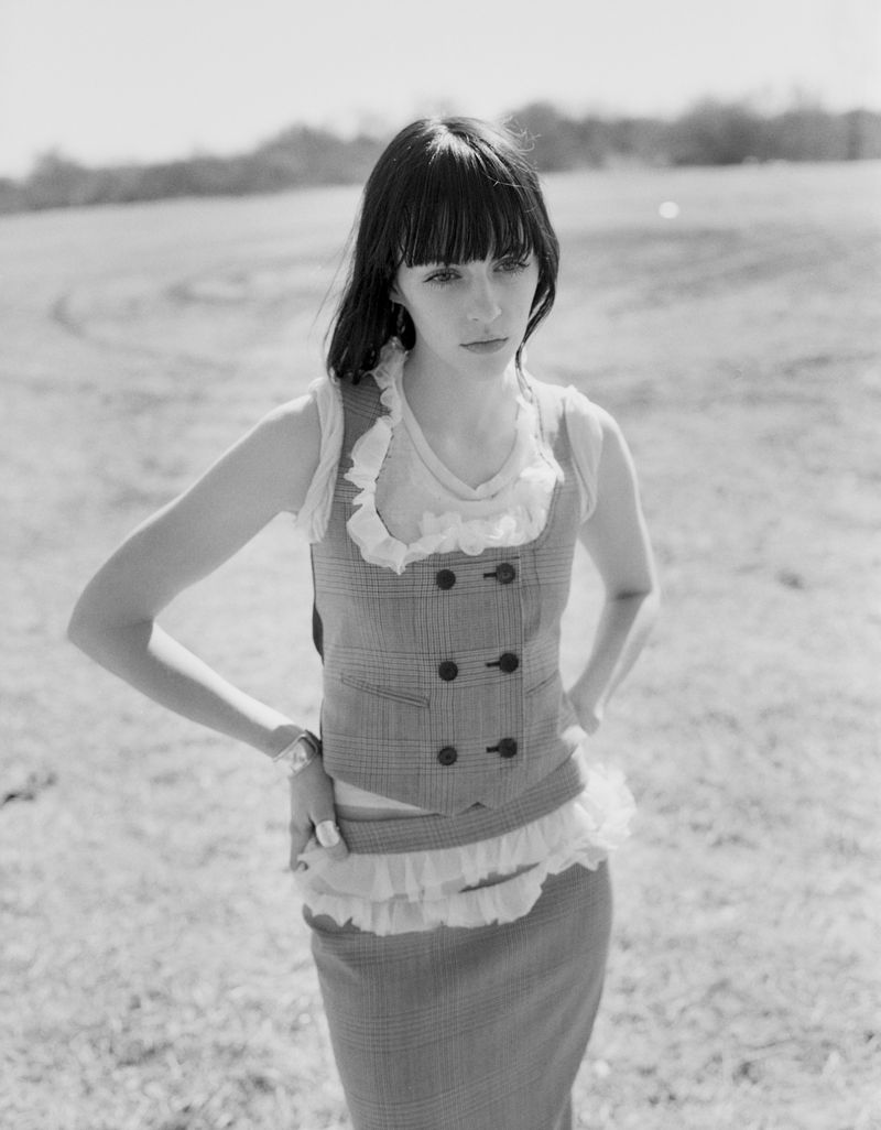 Model wearing a double-breasted patterned vest and matching skirt standing in an open grassy field.