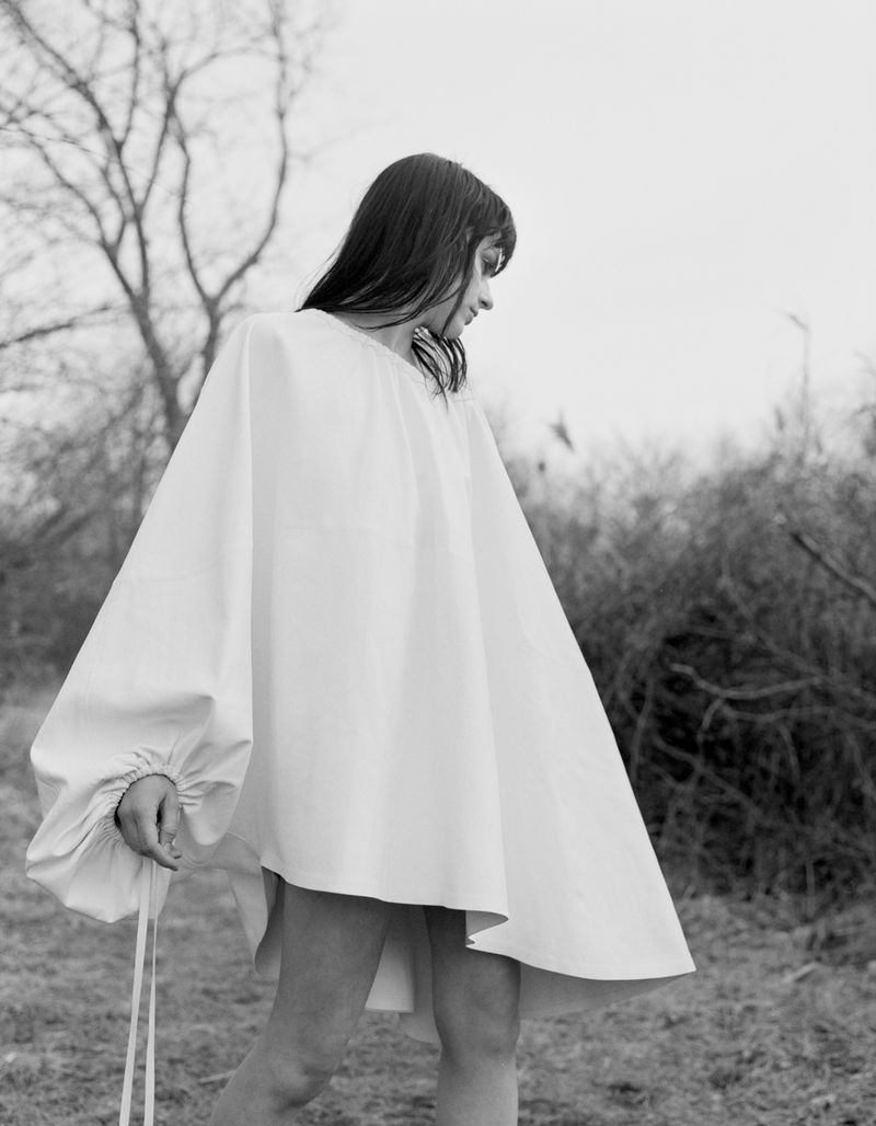 Woman in white poncho stands in field with bare trees against overcast sky; minimalist fashion and rural environment, monochrome portrait.