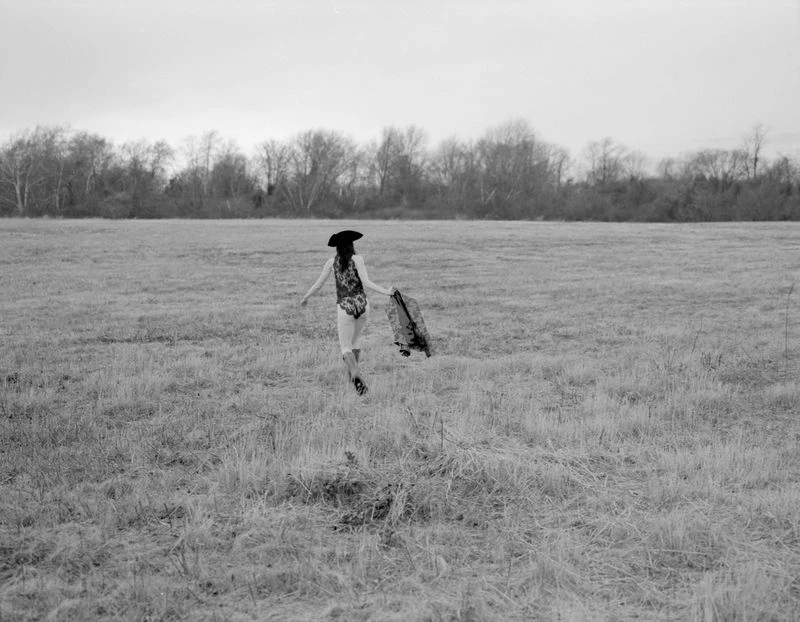 A person in a floral swimsuit and hat runs through a field with trees in the distance, captured in a black and white photograph.
