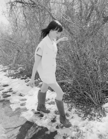 Monochrome fashion editorial image featuring a young woman walking near bare winter trees and patches of snow.