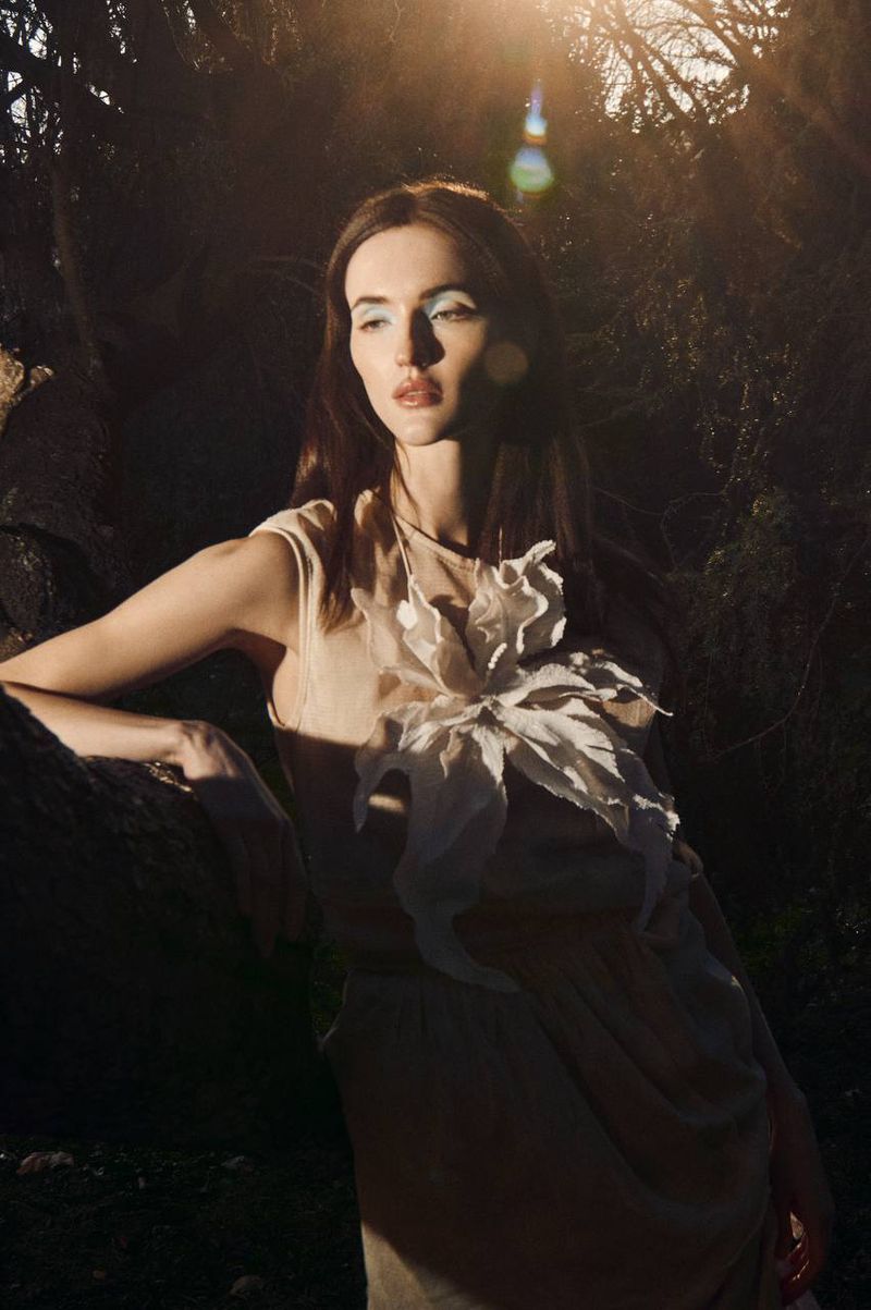 Brunette woman in natural light, wearing a light beige gown with a flower embellishment, posing in a forest environment.