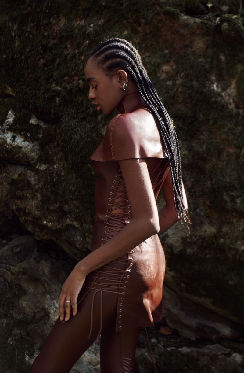Elegant woman with braids poses in a brown dress outdoors against a mossy rock backdrop, showcasing fashion and natural beauty.