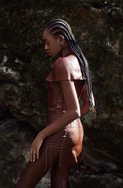 Elegant woman with braids poses in a brown dress outdoors against a mossy rock backdrop, showcasing fashion and natural beauty.