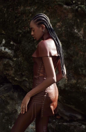 Elegant woman with braids poses in a brown dress outdoors against a mossy rock backdrop, showcasing fashion and natural beauty.