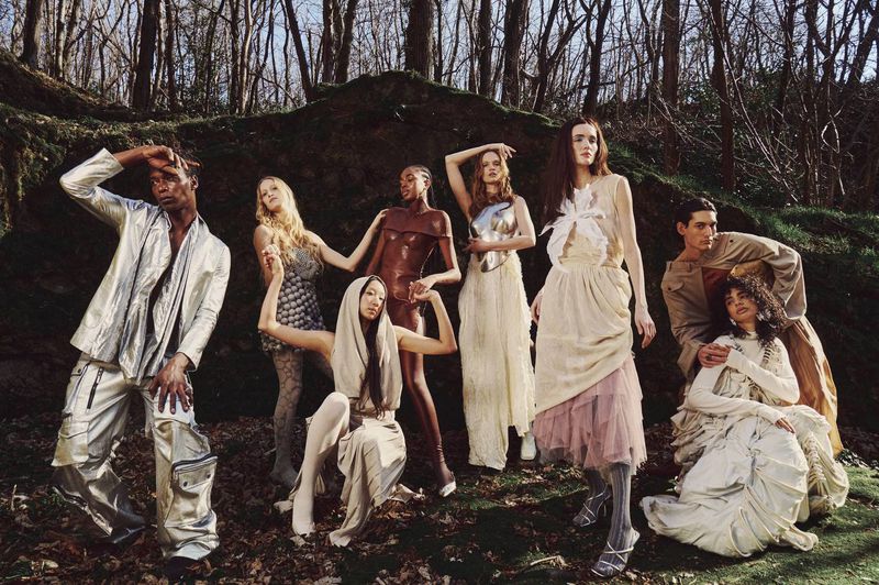 A group of eight fashion models pose in a wooded area, wearing a variety of light-colored and metallic outfits in an outdoor setting.