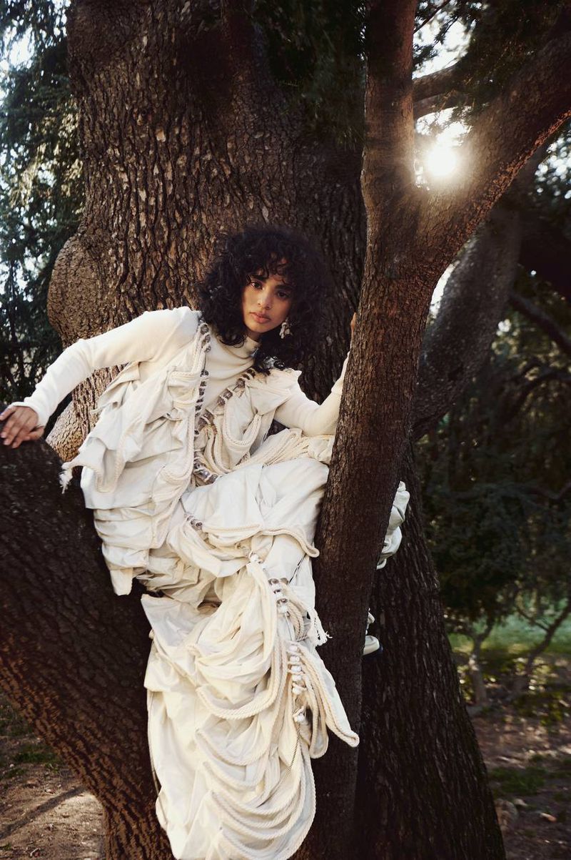 Model with dark curly hair wearing a white textured layered outfit perched in a large tree