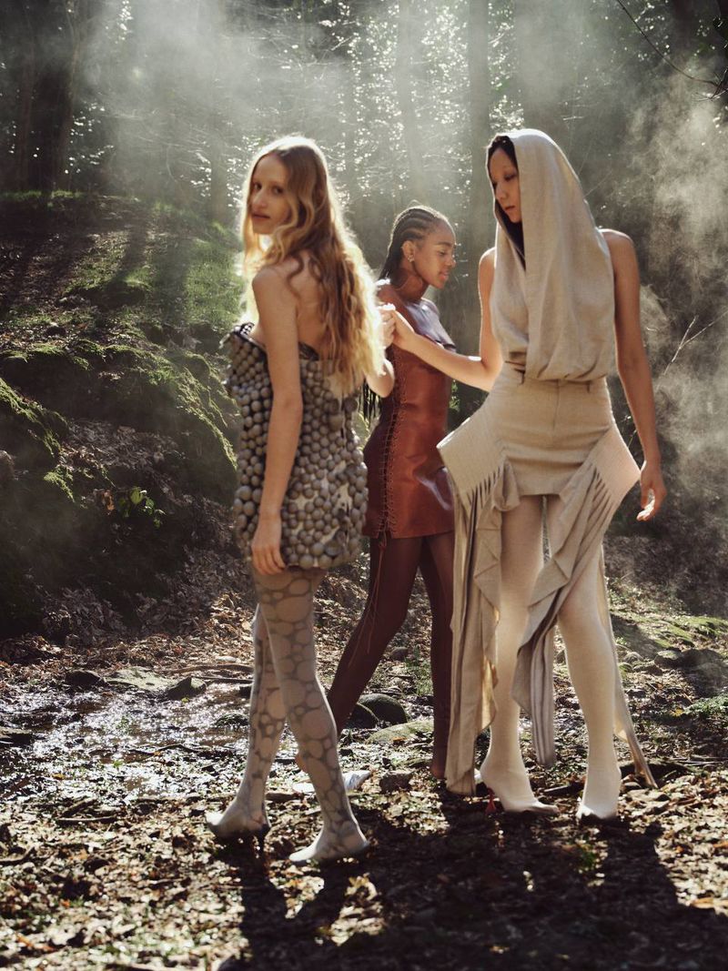 Three fashion models pose in a forest. One wears a strapless dress covered in round objects and patterned tights. A second wears a leather-look dress.