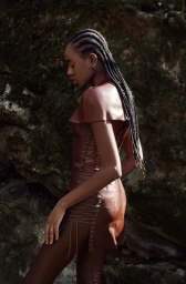 Black model with braided hair wearing a brown leather dress and matching leggings against rocky ground