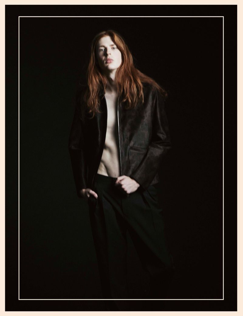 Portrait of a slender adult man with long red hair, wearing a dark leather jacket and black pants, standing in a dark photo studio.