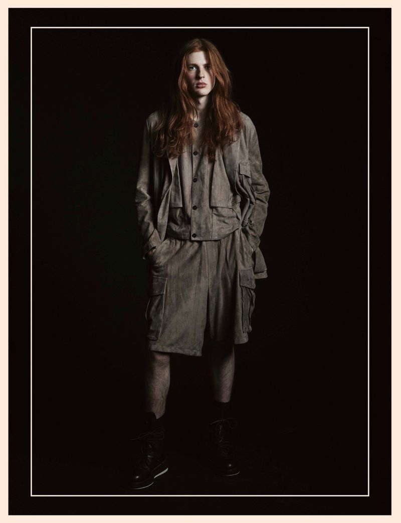 Fashion shot of a male model with long red hair in a beige corduroy two-piece outfit, posing in a photo studio with a dark background.