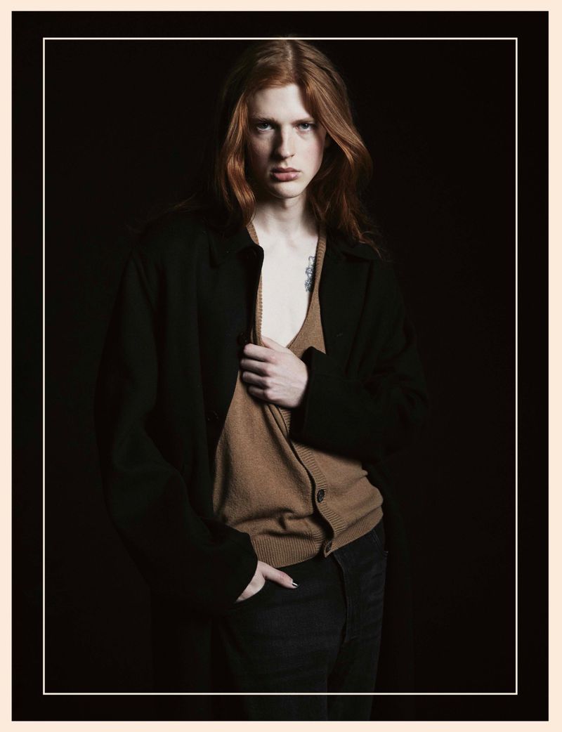 Portrait of a young, fair-skinned male model with long red hair wearing a black coat and tan cardigan against a dark background.