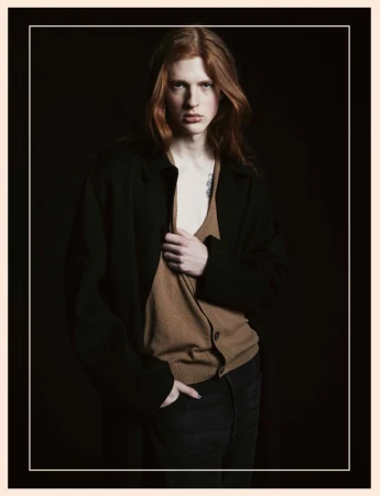 Portrait of a young, fair-skinned male model with long red hair wearing a black coat and tan cardigan against a dark background.