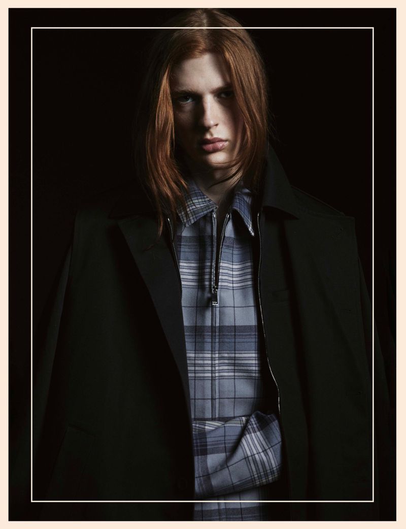 Portrait of a red-haired male adult wearing a blue plaid shirt and black overcoat against a dark backdrop in a studio setting.