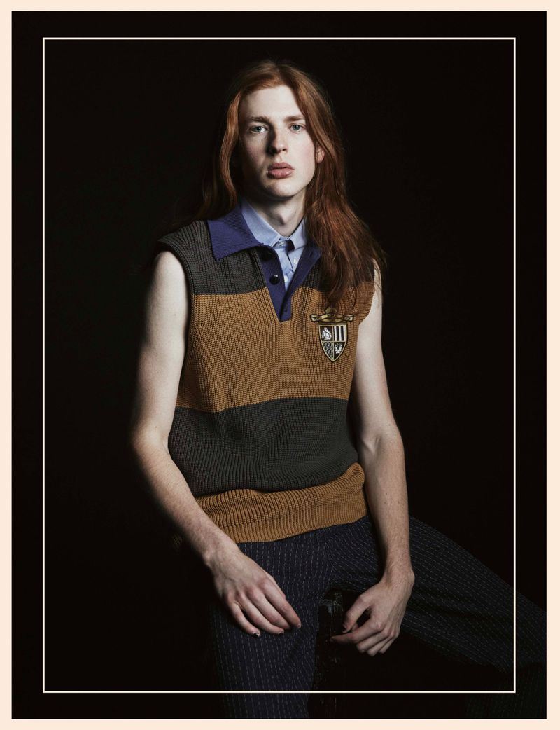 Studio portrait of a fair-skinned young adult male with long red hair, wearing a brown sleeveless knit sweater, in a dark indoor setting.