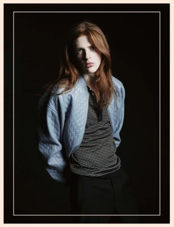 Model with long reddish hair wearing a light blue textured bomber jacket against a dark background