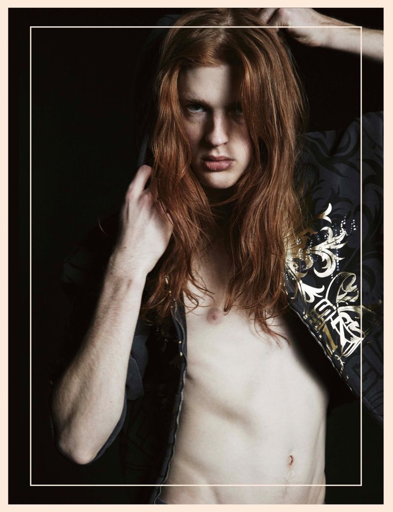 Portrait of a shirtless adult male with long red hair, dark jacket with gold accents, against a dark background in studio lighting.