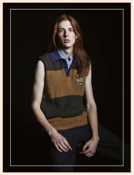 Long-haired male model wearing a striped sweater vest and collar shirt against a dark background
