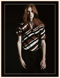 Model with long red hair wearing a striped knit polo and black trousers against a dark background for Numero Homem Brasil