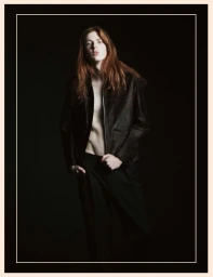 Model with long red hair wearing a dark unzipped jacket and black trousers against a dark background