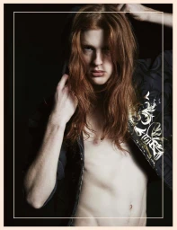 Portrait of a long-haired male model in an unzipped patterned jacket against a dark background.