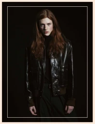 Model with long red hair wearing a dark leather bomber jacket against a black background inside a framed border