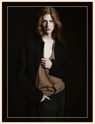Long-haired male model wearing a dark coat and brown V-neck cardigan against a black background with a white frame border.