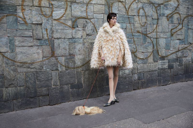 Stylish woman in a fluffy coat walks a golden-haired dog on a leash in an urban setting with a textured grey stone wall backdrop.