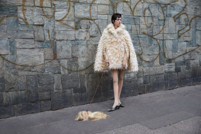 Stylish woman in a fluffy coat walks a golden-haired dog on a leash in an urban setting with a textured grey stone wall backdrop.