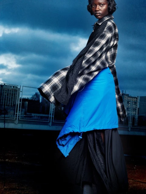 Fashion editorial portrait of a woman in a blue skirt and flannel shirt, posing against a cloudy sky and urban cityscape backdrop.