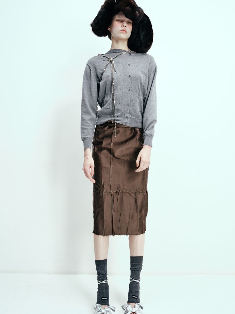 Fashion portrait of a single adult female wearing a gray cardigan, brown skirt, statement shoes and a large fur hat, against white backdrop.