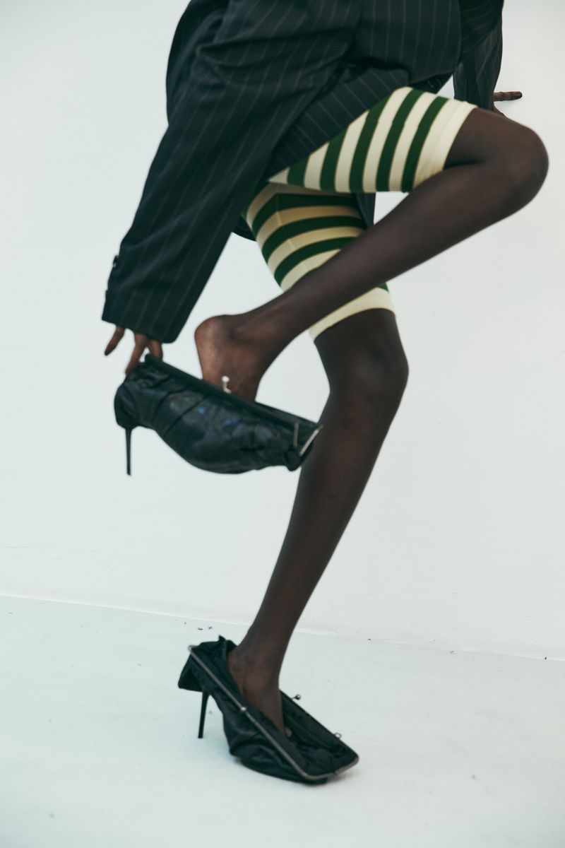 Fashion shot of a Black woman with legs and feet visible, stepping into black heels. She wears striped bike shorts and a pinstripe jacket.