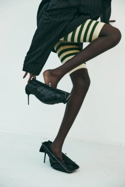 Fashion shot of a Black woman with legs and feet visible, stepping into black heels. She wears striped bike shorts and a pinstripe jacket.
