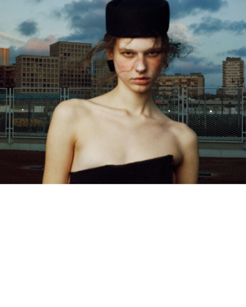 Model wearing a black pillbox hat and strapless top against an urban rooftop and skyline background.