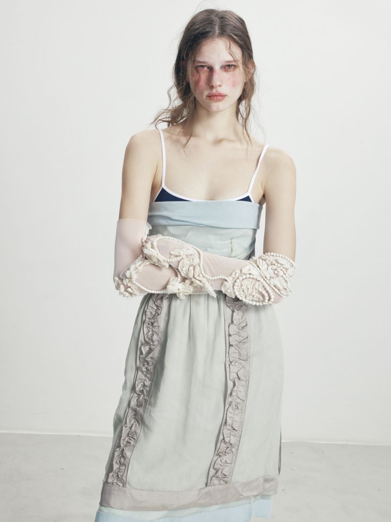 A young woman poses in a blue slip dress and lace gloves, standing in a photo studio with fair skin and light brown hair.
