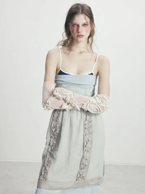 A young woman poses in a blue slip dress and lace gloves, standing in a photo studio with fair skin and light brown hair.