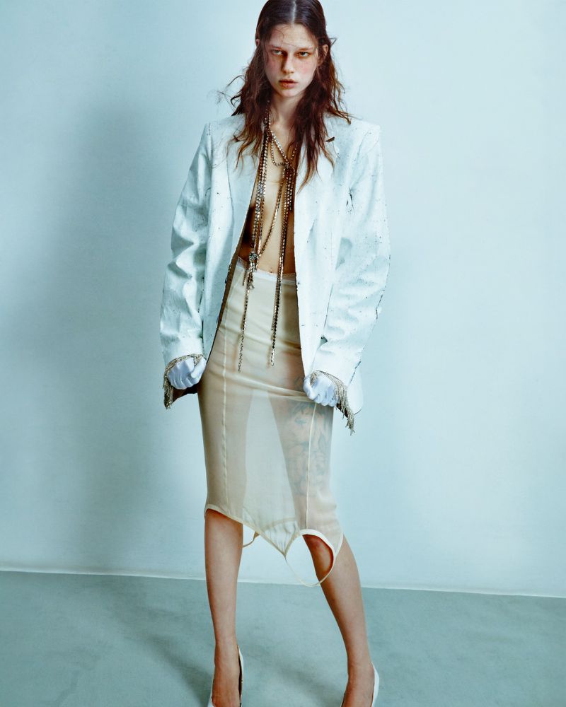 Model wearing a structured white blazer and sheer nude skirt standing against a pale grey studio background.