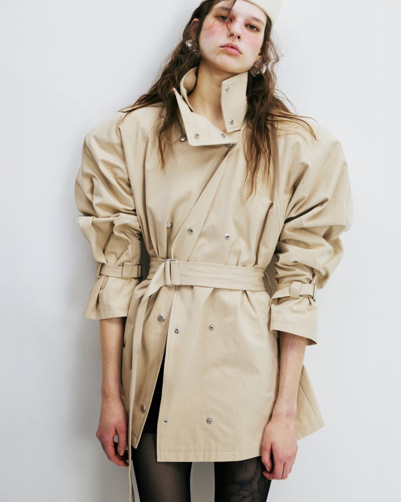 Fashionable woman in a beige trench coat with a belt, tulle tights, and a white hat poses in a studio with a white wall.