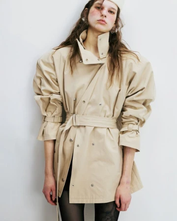Fashionable woman in a beige trench coat with a belt, tulle tights, and a white hat poses in a studio with a white wall.