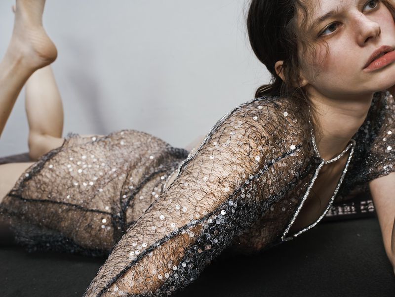 A young woman lies on a black surface wearing a sheer, shimmering dress with a beaded necklace against a neutral grey background.