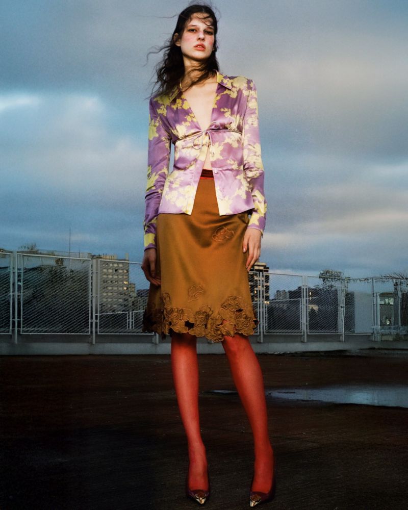 A fashionable young adult woman poses on a rooftop wearing a purple floral blouse, a brown skirt, red tights, and gold high heels.