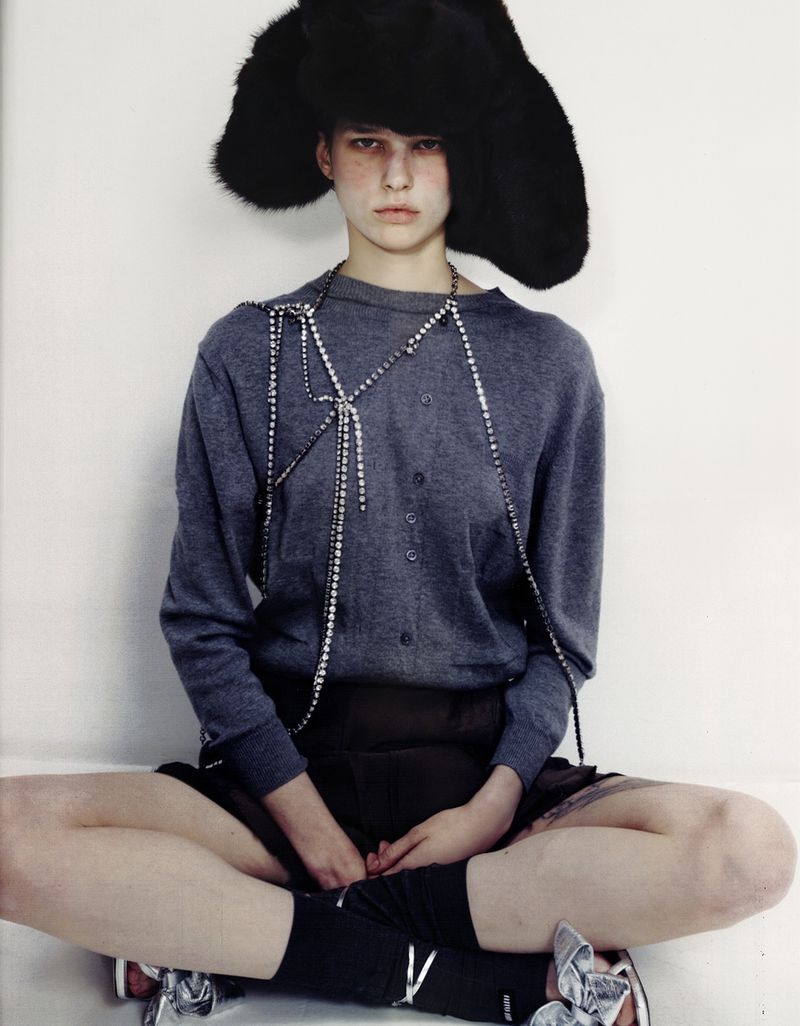Fashion shot of a person with black furry hat, grey sweater, and silver shoes sitting cross-legged against a white background in studio.