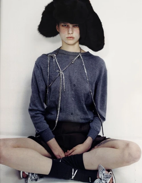 Fashion shot of a person with black furry hat, grey sweater, and silver shoes sitting cross-legged against a white background in studio.