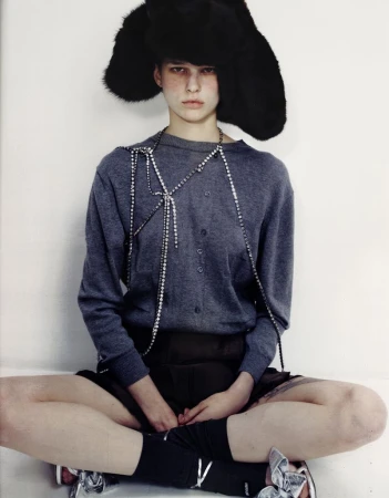 Fashion shot of a person with black furry hat, grey sweater, and silver shoes sitting cross-legged against a white background in studio.
