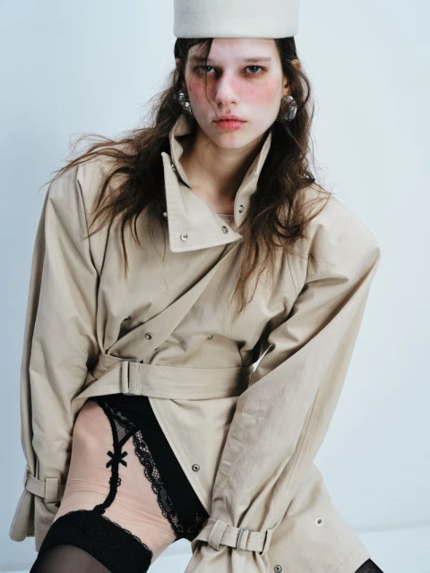 A young adult female model poses in a trench coat, hat, and lingerie against a white background for a studio fashion shoot.