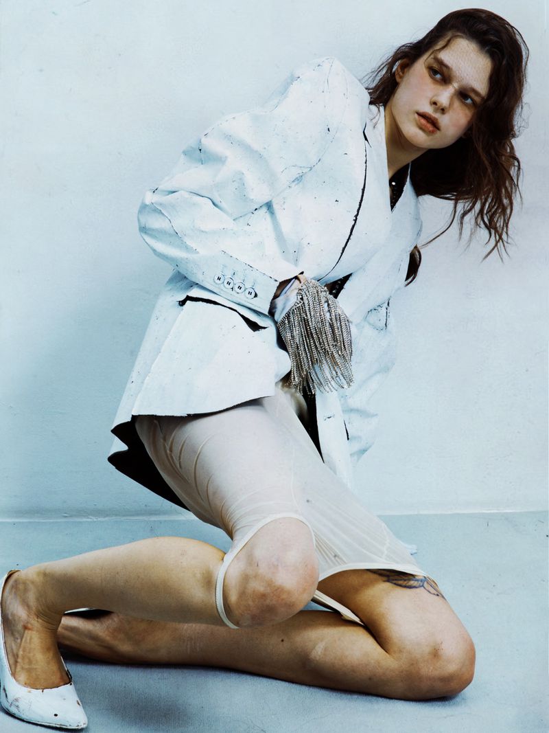A fashion photo of a young adult woman with light brown hair in white heels, a white coat, and white underwear kneeling in a photo studio.