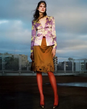 Model standing outdoors wearing a purple floral satin blouse and brown skirt against a cloudy sky.