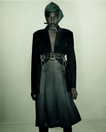 Model wearing a black blazer, grey pleated skirt, and green head covering against a neutral background