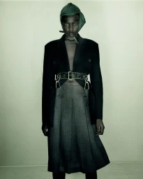 Model wearing a black blazer, grey pleated skirt, and green head covering against a neutral background